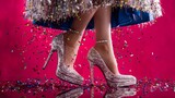 A pair of sparkling high-heeled shoes against a vibrant pink background. The shoes are adorned with shimmering sequins and are positioned on a surface scattered with multicolored confetti. 