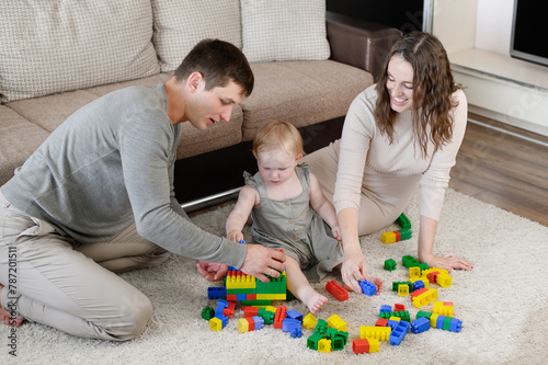 Happy family with child, sitting on floor at home, builds toy tower from multi-colored plastic blocks. Joyful child plays with mom, dad, spending family time together. Educational game for children