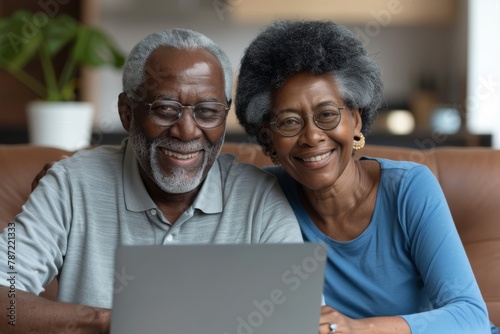 A happy black senior couple sits together at a laptop, looking into the camera and smiling, websurfing on internet with laptop at home and studying the laptop screen intently