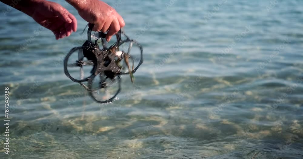A close-up of the hand of a man who takes the FPV drone out of the water in the ocean and removes algae from it and shakes it off the water. Drone crash, breakdown.