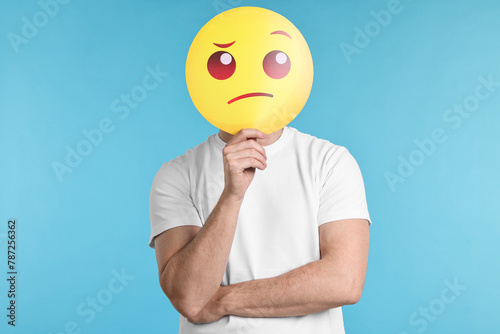 Man covering face with thinking emoticon on light blue background © New Africa