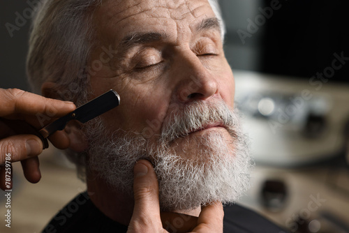 Professional barber shaving client's beard with blade in barbershop