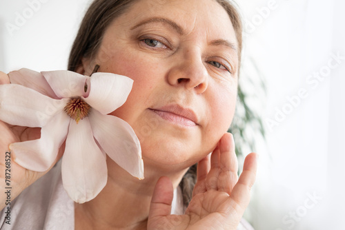 aging woman with magnolia flower looks herself in mirror, noticing wrinkles, changes in facial contour and sagging eyelids, cosmetic anti-aging procedures, natural process growing older