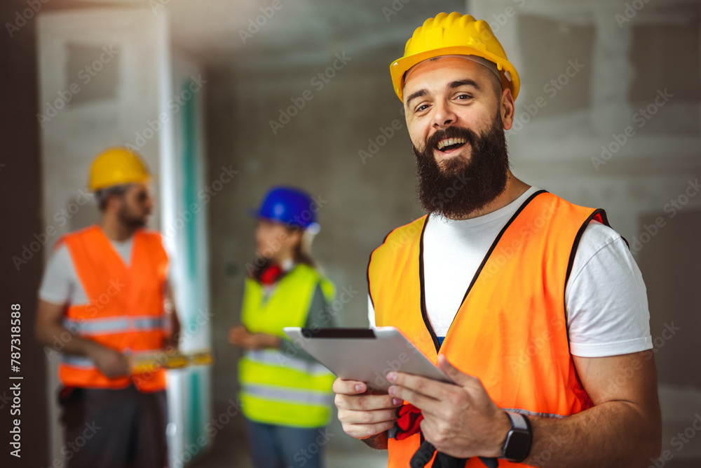 Construction site manager standing  wearing safety vest and helmet, holding a tablet.