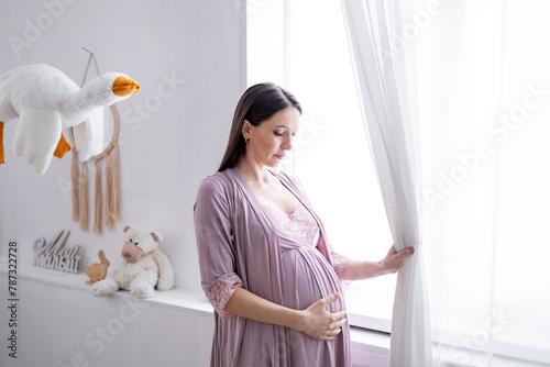 A pregnant woman strokes her big belly looking out the window in a bright children's bedroom by the crib. The expectant mother is waiting and preparing for the birth of a child