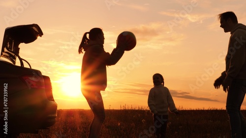 Family playing with ball near car at sunset, silhouettes. Father, mother, daughter throwing ball in circle, laughing, having fun joyful good time in field, creating pleasant unforgettable memories.