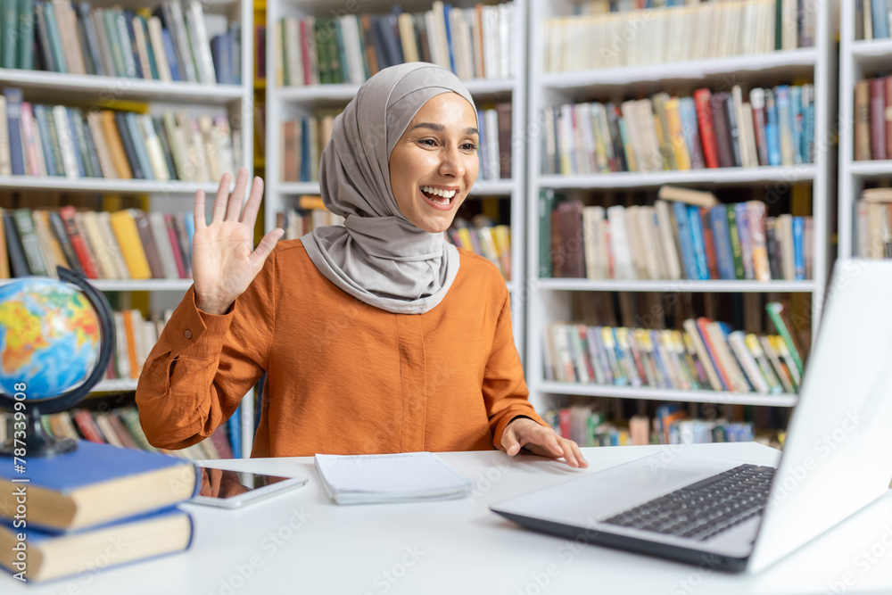 Joyful Muslim woman wearing a hijab waves at her laptop during a friendly video call, surrounded by books in a well-stocked library. Joyful Muslim woman wearing a hijab waves at her laptop during a friendly video call, surrounded by books in a well-stocked library.