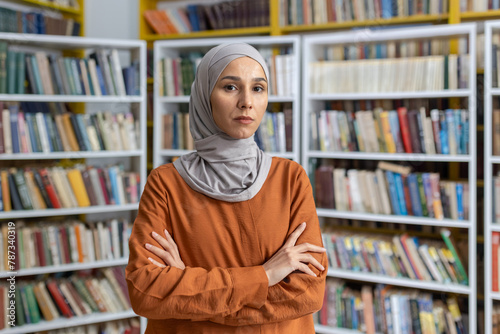 A confident Muslim woman in a hijab stands with her arms crossed in a library filled with books, portraying strength and intelligence.