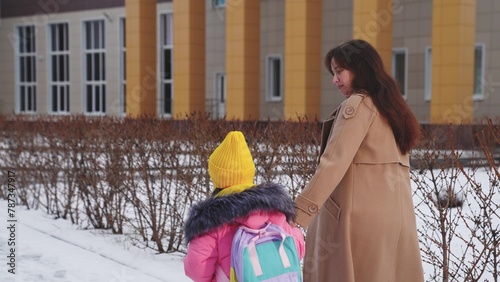 Happy family, mother holding chilt by hand leads school, schoolgirl with her mother walks through school yard by hand, Child Kid daughter walks with her mother school snow, winter school yard, mother