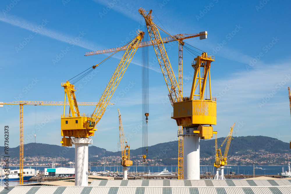 This image showcases several large yellow industrial cranes towering over a construction site with a clear blue sky This image showcases several large yellow industrial cranes towering over a construction site with a clear blue sky