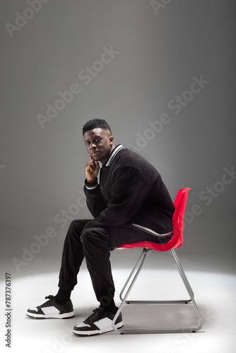A black man dressed in a suit is seated on a vibrant red chair against a neutral background.