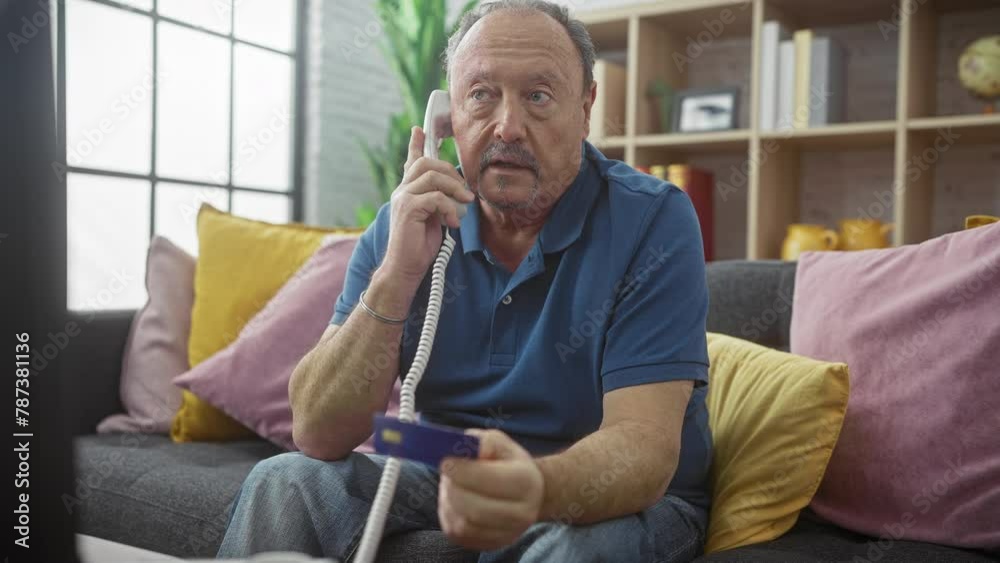 Mature man in a living room talking on the phone while holding a credit card, displaying a mix of concentration and concern. Mature man in a living room talking on the phone while holding a credit card, displaying a mix of concentration and concern.