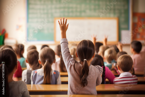 Schoolgirl raising her hand in the classroom. The girl is seen from the back, behind other school kids, some also with their hands raised and eager to answer.