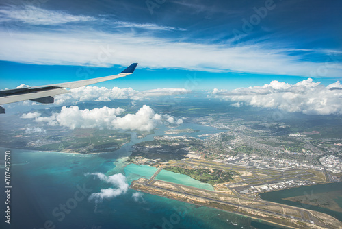 "Above the Horizons: An aerial vista captures Oahu, Hawaii, with the airport below, framed by billowing clouds, a vast expanse of blue sky, and the distant shimmer of ocean waters"