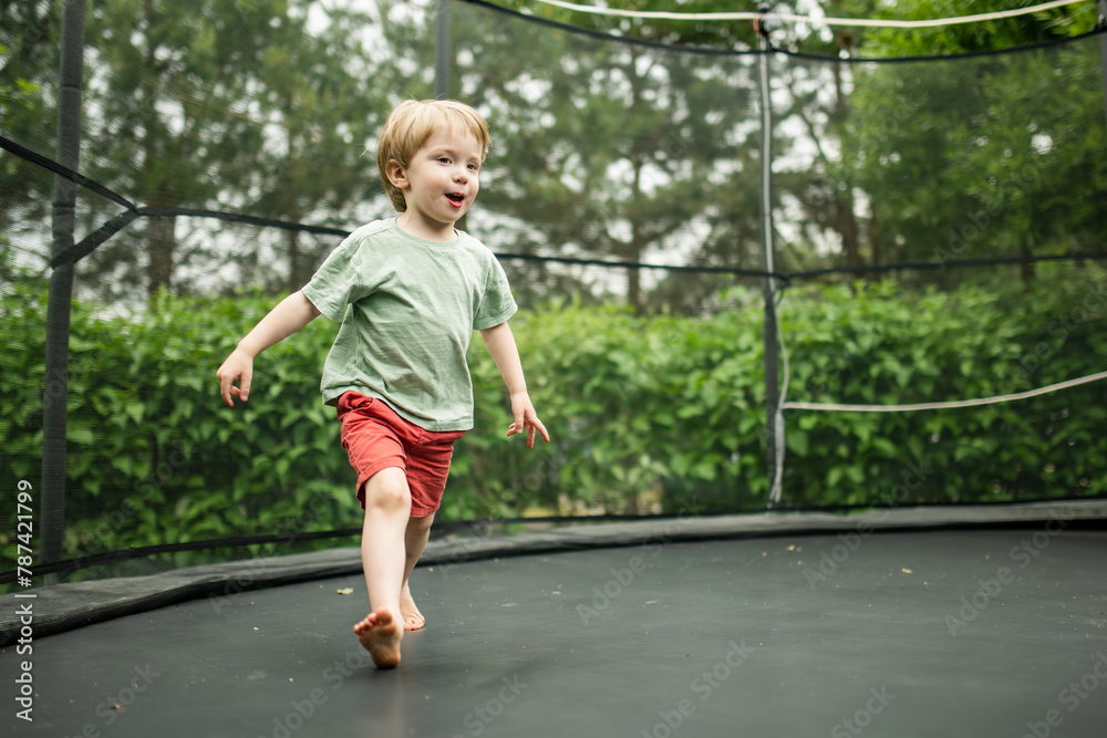 Cute little boy jumping on a trampoline in a backyard on warm and sunny summer day. Sports and exercises for children. Summer outdoor activities. Cute little boy jumping on a trampoline in a backyard on warm and sunny summer day. Sports and exercises for children. Summer outdoor activities.