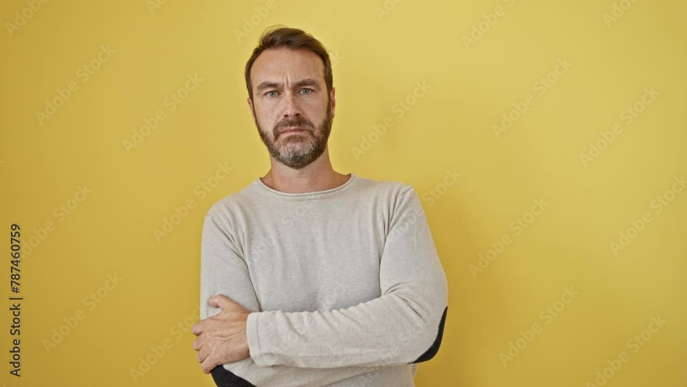 Skeptical middle-aged hispanic man, disapproving expression on his face, nervously standing with crossed arms, isolated on a yellow background Skeptical middle-aged hispanic man, disapproving expression on his face, nervously standing with crossed arms, isolated on a yellow background