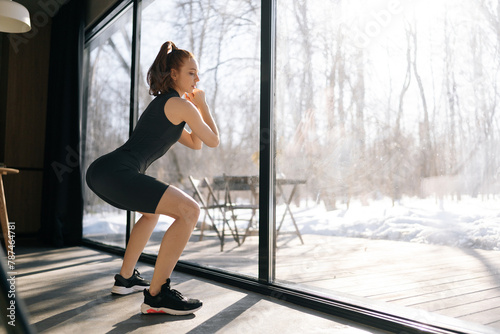 Back view of slim fit young woman in activewear making squats training at home. Athletic fit female doing fitness aerobic exercises for booty in room standing by window on sunny morning.