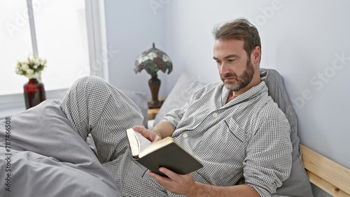 Middle-aged bearded man in pajamas reading a book while relaxing in a well-lit bedroom interior. photo