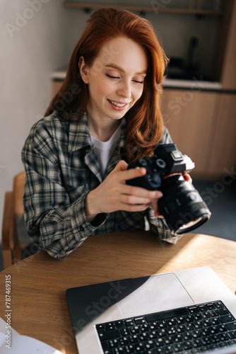 Vertical portrait of young female photographer holding modern digital camera looking at screen choosing photos for editing while sitting in front of laptop computer at home office workplace.