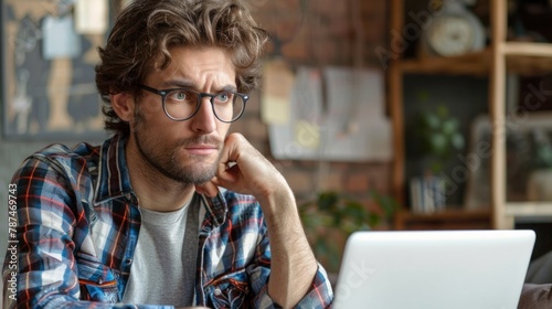 Man in his thirties dressed casually infront of his laptop with a pondering look on his face. He looks like he is trying to solve a problem photo