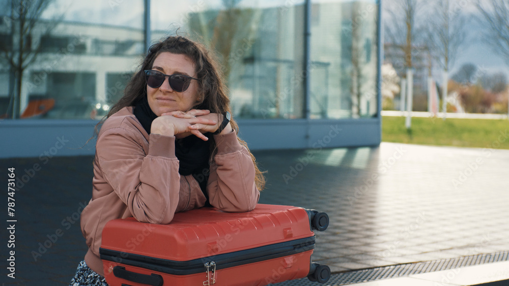 Fototapeta premium A young woman with a red suitcase is waiting for a transport, a bus outdoors. The wind blows the hair. Urban lifestyle
