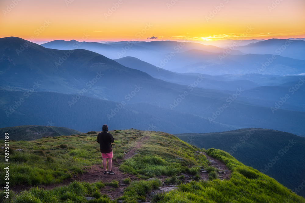 Obraz premium Man on the top of mountain Goverla, Ukraine. Hill with green grass and beautiful mountain valley at sunset in summer. Landscape with sporty man, hils in fog, orange sky. Hiking. Spring in Europe