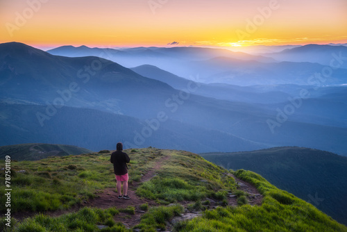 Man on the top of mountain Goverla, Ukraine. Hill with green grass and beautiful mountain valley at sunset in summer. Landscape with sporty man, hils in fog, orange sky. Hiking. Spring in Europe