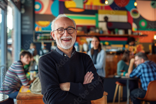 A cheerful elderly man with glasses stands with crossed arms in a vibrant cafe, his warm smile reflecting a contented life surrounded by patrons.