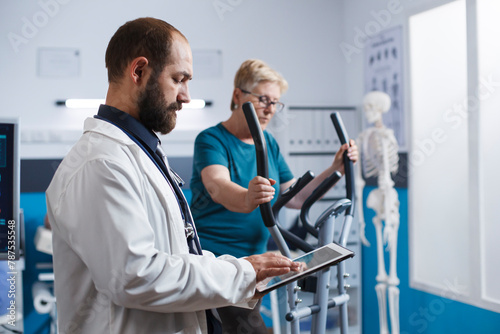 Portrait of doctor holding tablet while elderly patient uses electronic bicycle for physical rehabilitation. Male medical physician in lab coat using digital device for medical physiotherapy research.