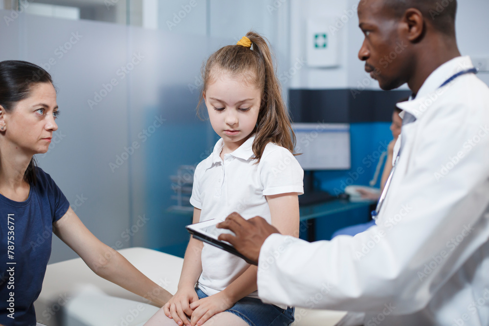 Obraz premium Black man wearing lab coat consults with girl patient and mother in clinic office. African American physician using digital tablet while providing medical advice to caucasian woman and her daughter.