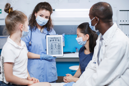 In clinic office  orthopedic doctor and nurse discuss the best treatment choices for the patient. Skeletal information is displayed on a tablet by a doctor wearing blue scrubs and a face mask.
