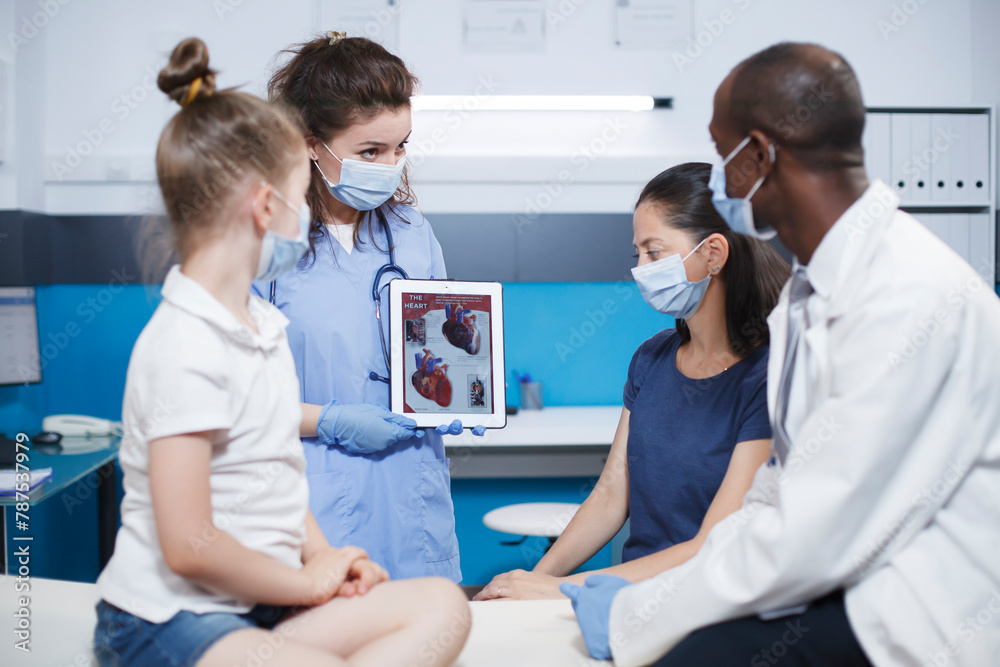 Fototapeta premium Black doctor shows cardiology image on digital tablet, pediatrician explains cardiovascular diagnosis to child and parent. Heart anatomy picture and heart disease problems presented by pediatrician.