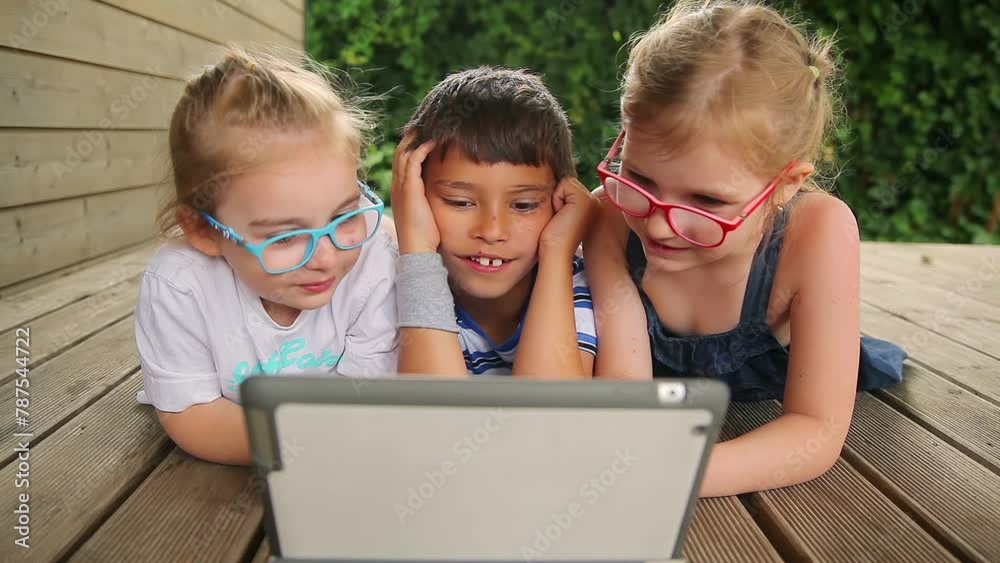 Three children lie on the veranda and look at the tablet. High quality 4k footage Three children lie on the veranda and look at the tablet. High quality 4k footage