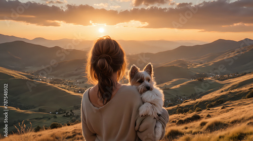 A woman with her hair tied back holds a small dog, both looking out over a stunning, golden-lit landscape of rolling hills at sunset