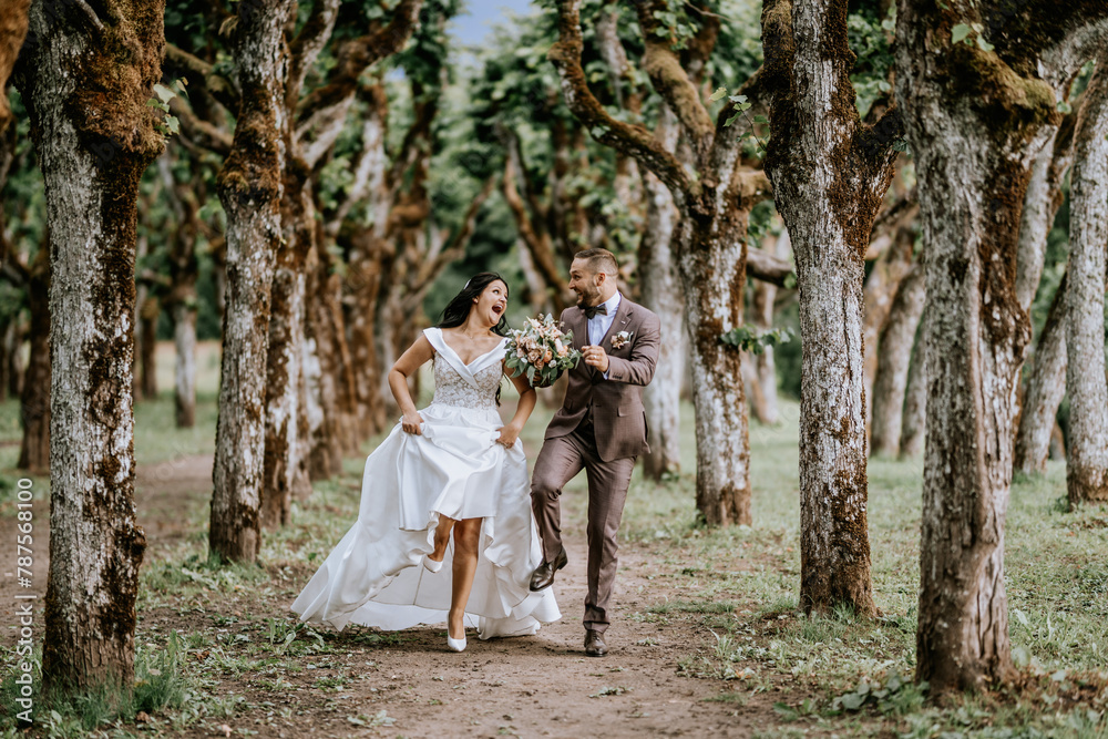 Valmiera, Latvia- July 28, 2024 - A joyful bride and groom run hand in hand down a path lined with knotty trees, sharing a laugh in a candid moment on their wedding day
