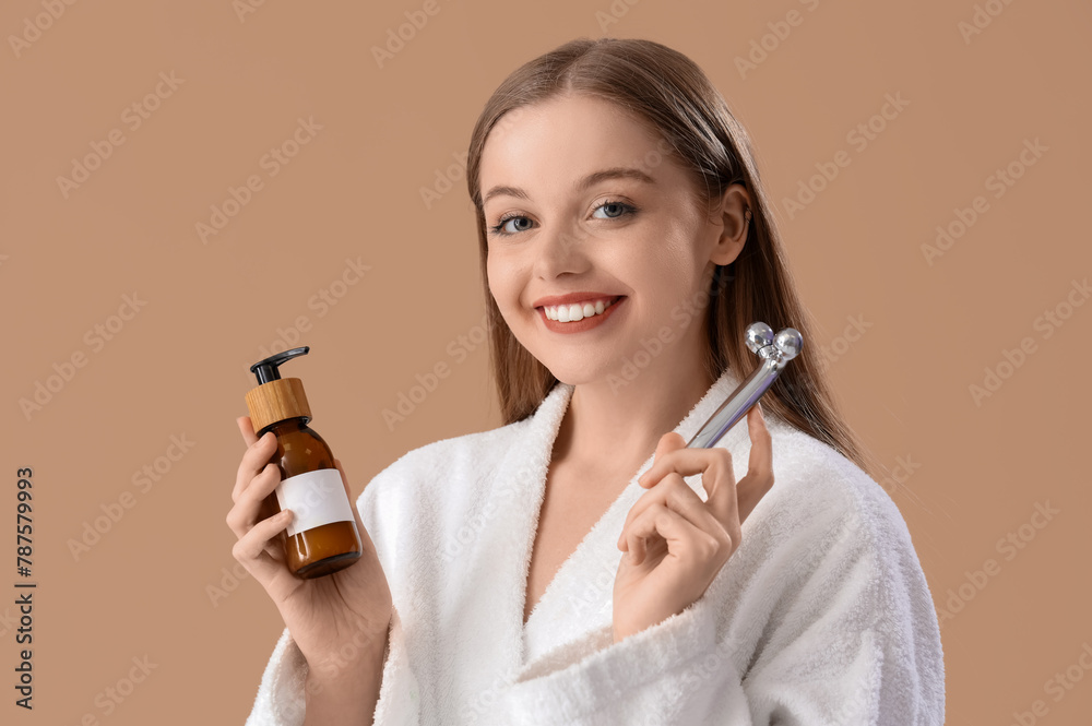 Young woman in bathrobe with facial massage tool and bottle of cream on beige background, closeup Young woman in bathrobe with facial massage tool and bottle of cream on beige background, closeup