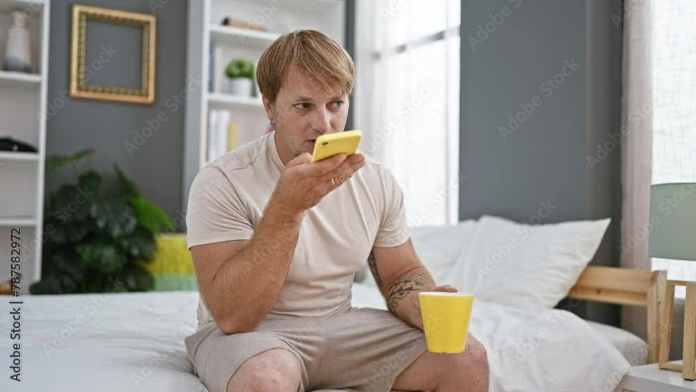 A young bearded man using a smartphone while sitting on a bed in a modern bedroom interior, holding a yellow cup. A young bearded man using a smartphone while sitting on a bed in a modern bedroom interior, holding a yellow cup.
