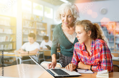 Friendly female teacher helps schoolgirl find information in laptop in the school library photo