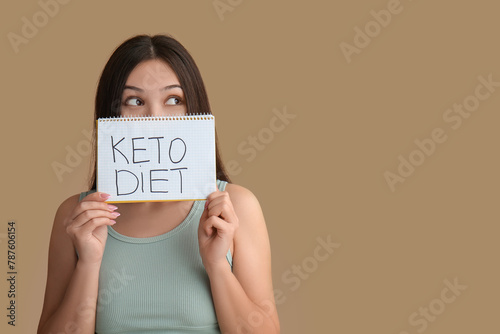 Beautiful young thoughtful Asian woman holding notebook with text KETO DIET on brown background