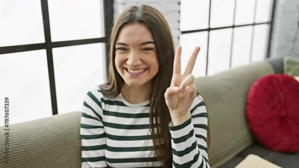 Young hispanic woman sitting on the sofa smiling with happy face winking at the camera doing victory sign. number two. at home Young hispanic woman sitting on the sofa smiling with happy face winking at the camera doing victory sign. number two. at home