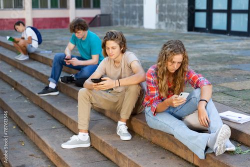 Teens with smartphones sitting on stairs near school building.
