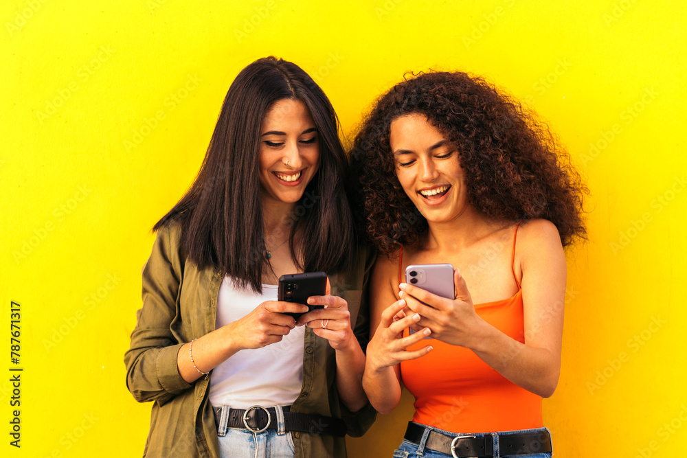 afro and caucasian women watching mobile leaning on a yellow wall afro and caucasian women watching mobile leaning on a yellow wall
