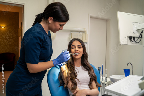 Young woman with dental braces having in dental office   wide shot  open mouth  female dentist using mirror for checkup 