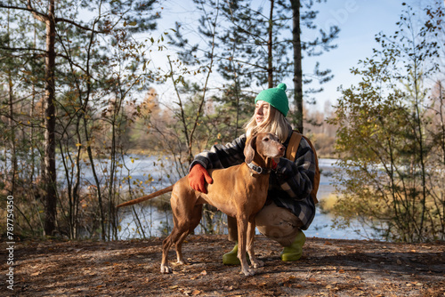 Middle aged woman petting Hungarian Vizsla dog walking in nature autumn park. Female travelling spending time with pet friend on weekend sitting on haunches. Travel, tourism with dog, wanderlust.