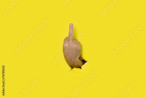 Hand of man making insulting gesture with middle finger through a hole in yellow paper background. Space for text.