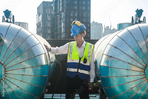 Engineer with safety gear inspects large industrial water tanks on a city rooftop against a skyline backdrop.