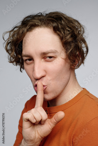 Vertical closeup studio portrait of sly young Caucasian man with curly hair showing be quiet or hush gesture photo