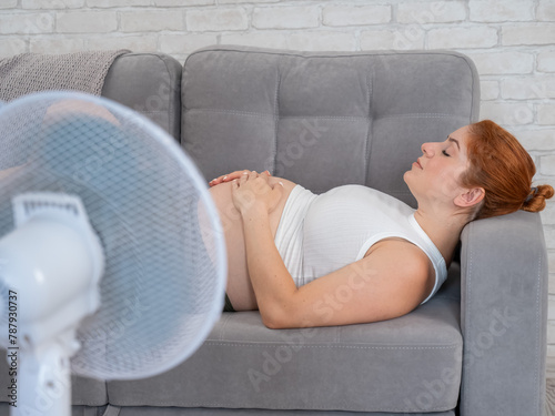 Pregnant woman suffers from heat and lies on the sofa near the electric fan.  © Михаил Решетников