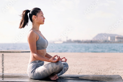 young asian woman in sportswear doing meditation by the sea on a yoga mat, concept of mental relaxation and healthy lifestyle, copy space for text