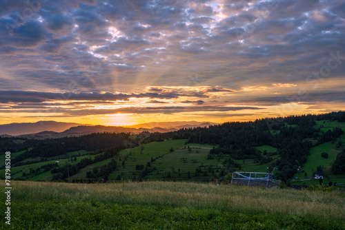 Sunrise over mountains. Cloudy sky with sunrays, green meadow in foreground in summer. © Marcin Mucharski